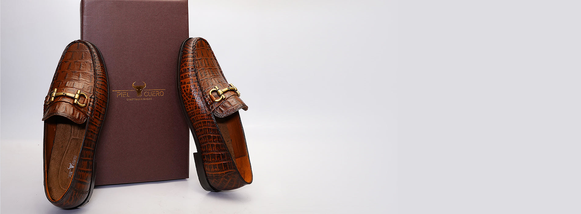 Premium brown leather loafers with crocodile texture and gold buckle detail, handmade by Piel Cuero, displayed with branded shoe box.